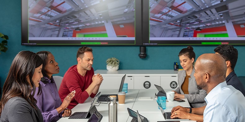 Several people at a conference table in front of TV monitors displaying a facility model 